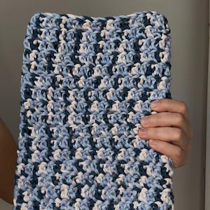 Peut inclure: Une pochette rectangulaire en crochet présentant un motif de fil panaché dans des tons bleu marine, bleu clair et crème. L'accessoire textile est réalisé avec un point dense et uniforme, créant une surface texturée sur tout l'objet.