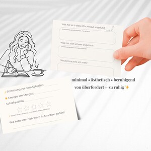 May include: Two cream-colored journaling cards with prompts for self-reflection. One card is held by a hand, the other is on a surface with a line drawing of a person writing. Text includes "minimal", "&auml;sthetisch", and "beruhigend".