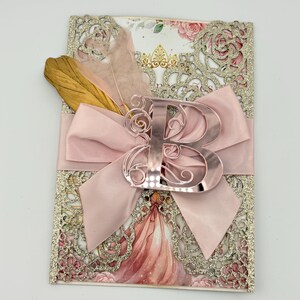 May include: A pink invitation card with a decorative laser-cut design and a large pink satin bow. The card features a metallic letter "B" and is adorned with gold and pink feathers. The background includes a floral illustration.