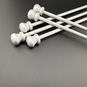 May include: A collection of silver-colored curtain rods with decorative finials. The rods are arranged diagonally across a dark background. The finials have a rounded, bulbous shape with a detailed design. The rods appear to be made of metal.