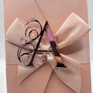 May include: A blush pink invitation card with a satin ribbon bow and a decorative rose gold letter "A" accent. The card is displayed on a clear stand, suitable for weddings or special events.
