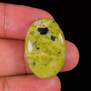 Peut inclure: Pierre précieuse ovale avec une couleur de base vert clair et des inclusions noires. La pierre polie a une surface lisse et un léger éclat. La pierre est tenue entre les doigts sur fond noir.