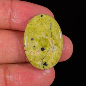 Puede incluir: Una gema ovalada de color verde claro, salpicada de manchas negras y finas líneas negras. La superficie pulida refleja la luz, mostrando los patrones y texturas naturales de la piedra. La gema se sostiene entre los dedos sobre un fondo negro.
