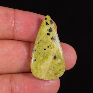 Peut inclure: Une pierre précieuse en forme de goutte d'eau, de couleur vert clair avec de petites inclusions noires. La pierre polie a une surface lisse et est tenue dans la main d'une personne sur un fond noir. La pierre est probablement utilisée pour la joaillerie.