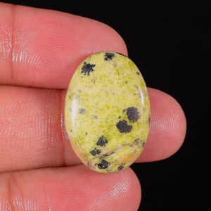 Peut inclure: Une pierre précieuse ovale de couleur jaune-vert clair, parsemée de taches gris foncé et noires. La surface polie reflète la lumière, révélant les motifs naturels. La pierre est tenue entre les doigts sur fond noir.