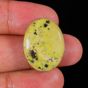 Puede incluir: Gema ovalada de color amarillo verdoso pálido, salpicada de inclusiones negras oscuras. La superficie pulida refleja la luz, mostrando los patrones naturales de la piedra. La gema se sostiene entre los dedos sobre un fondo negro.