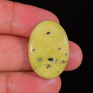Peut inclure: Pierre précieuse ovale de couleur jaune pâle, parsemée d'inclusions noires. La surface polie reflète la lumière, soulignant la texture lisse de la pierre. Le bijou est tenu dans une main sur fond noir.