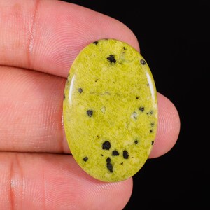 Peut inclure: Pierre précieuse ovale, de couleur vert clair, parsemée d'inclusions noires. La surface polie reflète la lumière, soulignant la texture naturelle et les variations de couleur de la pierre. La pierre est tenue entre les doigts sur fond noir.
