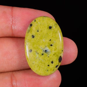 Puede incluir: Gema ovalada de color amarillo verdoso vibrante, salpicada de pequeñas inclusiones negras. La superficie pulida refleja la luz, resaltando los patrones naturales de la piedra. La gema se sostiene entre los dedos sobre un fondo negro.