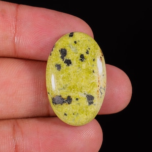 Puede incluir: Gema ovalada de color amarillo verdoso vibrante, salpicada de inclusiones grises oscuras y negras. La superficie pulida refleja la luz, mostrando los patrones naturales de la piedra. Una vista de cerca resalta la textura y las variaciones de color únicas de la gema.