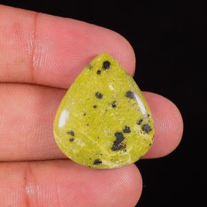 Puede incluir: Una gema en forma de lágrima, de color verde claro, con inclusiones negras dispersas. La piedra pulida se sostiene entre los dedos sobre un fondo oscuro, resaltando su superficie lisa y sus patrones naturales. Esta gema se utiliza probablemente para joyería.