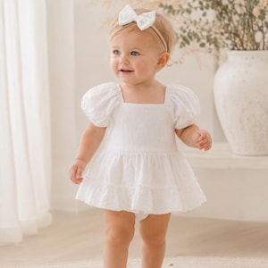 May include: A white eyelet romper featuring short puff sleeves, a square neckline, and a tiered ruffled skirt. The garment is adorned with a subtle floral pattern throughout the fabric. A matching white fabric bow headband is worn on the head.