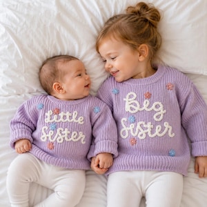 May include: Two lavender knit sweaters featuring white embroidered text and small floral accents. One sweater displays the words little sister, while the other displays big sister. Both garments have long sleeves, ribbed crew necklines, and cuffs.