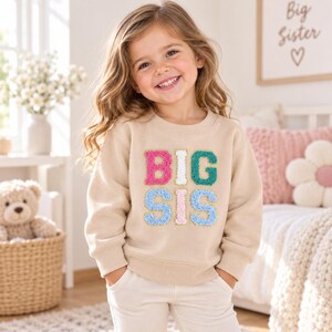 Può includere: Una felpa beige a maniche lunghe con la scritta BIG SIS in lettere stile varsity multicolori, testurizzate e soffici. Le lettere sono disposte su due righe nei colori rosa, bianco, verde acqua e azzurro, ciascuna con un bordo dorato.