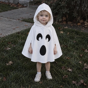 May include: A white hooded poncho featuring a black ghost face graphic with two arched eyes and an oval mouth. The garment has a loose, flared silhouette with arm openings on the sides, designed to be worn as a costume over everyday clothing.