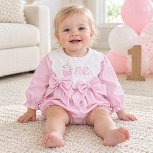 May include: A light pink long-sleeved romper with a white scalloped collar featuring an embroidered cupcake and the word one. The garment includes two large pink bows on the front, gathered cuffs, and a pinstriped fabric texture throughout.