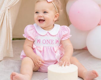 May include: A light pink and white pinstriped bubble romper with a white Peter Pan collar, short puffed sleeves, and pink ribbon bows at the waist. The bodice features smocking with the embroidered pink text ONE flanked by two pink bows.