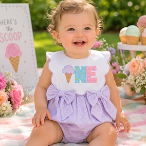 Può includere: Una tutina in seersucker bianco e viola chiaro con scollo smerlato e due grandi fiocchi in vita. Il corpetto presenta un grafico ricamato a forma di cono gelato e la parola ONE in lettere blu e rosa con granelli colorati.
