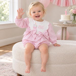 May include: A light pink long-sleeved romper featuring a white scalloped bib collar with an embroidered cupcake graphic and the word one. The garment includes two large pink bows at the waist and gathered cuffs on the sleeves, creating a soft texture.