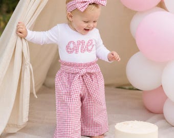 Könnte beinhalten: Ein langärmeliges weißes Oberteil mit dem Wort one in rosa Vichy-Karo-Muster, kombiniert mit einer passenden hoch taillierten Hose mit Bindegürtel und einem dazu passenden Haarband in rosa Vichy-Karo auf einem Kleinkind.