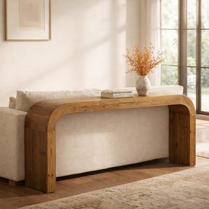 May include: A wooden console table with a curved design. The table is a light brown color and has a smooth surface. It is placed behind a beige sofa. A vase with dried flowers and a stack of books are on the table.