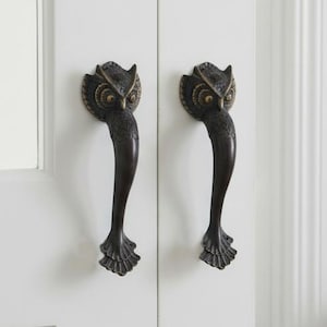 May include: Pair of bronze-toned owl-shaped door handles mounted on a white door. The handles have a detailed owl head design at the top and a curved handle. The door is part of a white interior with a table and vase in the background.