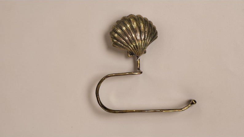 May include: A brass-coloured metal toilet roll holder with a seashell design. The holder features a detailed, fan-shaped shell at the top. The curved arm extends to hold a roll of toilet paper.