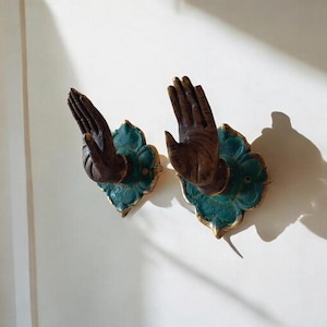 Könnte beinhalten: Zwei dekorative Wandhaken in Form von Händen, montiert auf teal- und goldfarbenen blütenförmigen Rückwänden. Die Hände sind dunkelbraun und die Haken sind an einer weißen Wand befestigt.