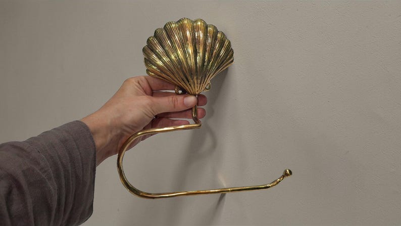 May include: A gold-coloured seashell-shaped toilet paper holder. The holder is made of metal and has a curved arm to hold the roll. The seashell design is detailed with ridges and a textured surface. The holder is mounted on a wall.