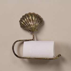 May include: A brass toilet roll holder with a seashell design. The holder has a detailed shell shape at the top and a curved bar to hold the toilet paper roll. It is mounted on a neutral-toned wall.