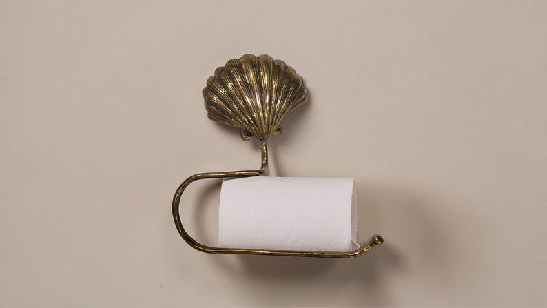 May include: A brass toilet roll holder with a seashell design. The holder has a detailed shell shape at the top and a curved bar to hold the toilet paper roll. It is mounted on a neutral-toned wall.