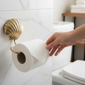 May include: A decorative gold-coloured seashell toilet roll holder fixed to a white marble tile wall. The holder has a curved arm to hold the roll of white toilet paper. The bathroom setting includes a toilet and a small wooden shelf.