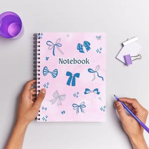 May include: Spiral-bound notebook with a pink cover featuring blue bow illustrations and the word "Notebook". The notebook is held by hands, with a purple pen and cup nearby. A paper clip with a purple handle is also visible.