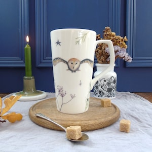 May include: A white ceramic mug with a brown owl design, flying with wings outstretched, against a background of white stars, a sprig of greenery, and a few small flowers. The mug has a black handle and is sitting on a wooden surface.