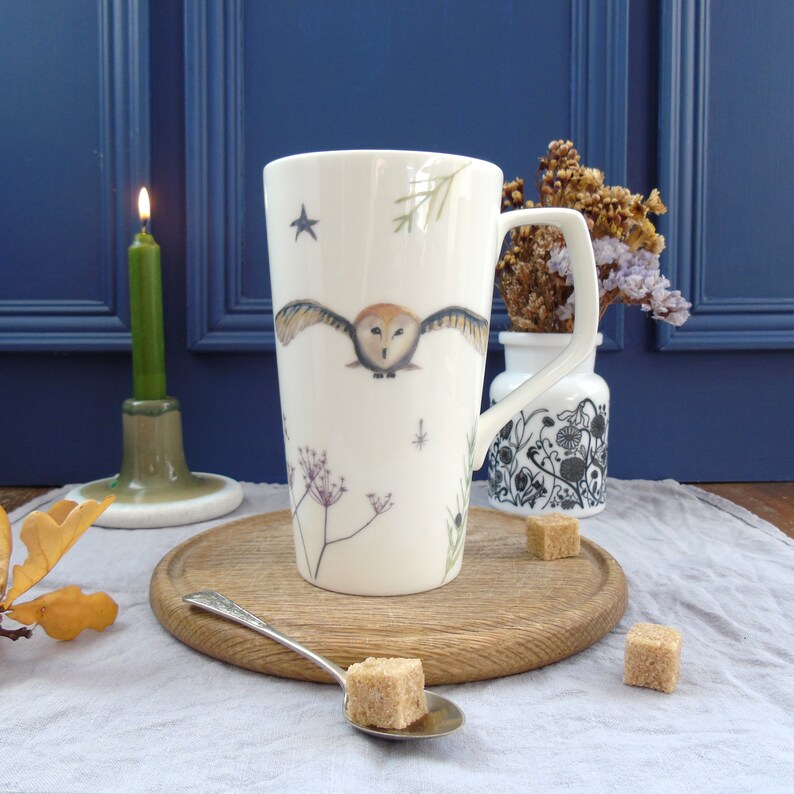 May include: A white ceramic mug with a brown owl design, flying with wings outstretched, against a background of white stars, a sprig of greenery, and a few small flowers. The mug has a black handle and is sitting on a wooden surface.