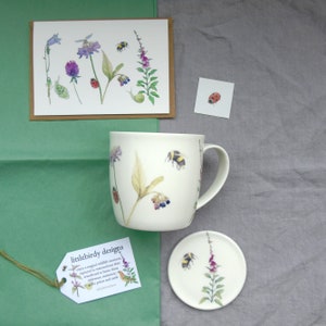 May include: A white ceramic mug with a bee and flower design. The mug is accompanied by a matching coaster and a card with the text "littlebirdy designs".