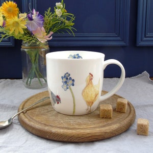 May include: White ceramic mug with a yellow chicken, a gray chicken, and blue forget-me-not flowers. The mug has a handle and is sitting on a wooden coaster.