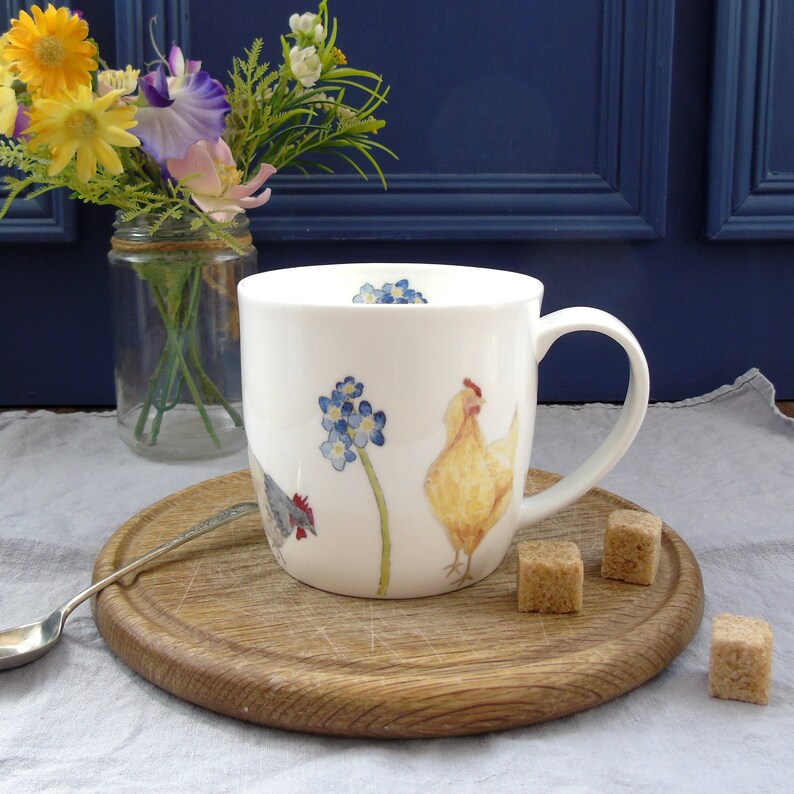 May include: White ceramic mug with a yellow chicken, a gray chicken, and blue forget-me-not flowers. The mug has a handle and is sitting on a wooden coaster.