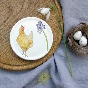 May include: A white ceramic plate with a hand-painted design of a yellow chicken and blue forget-me-not flowers. The plate is on a wooden surface.
