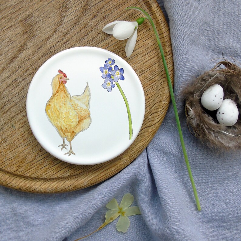 May include: A white ceramic plate with a hand-painted design of a yellow chicken and blue forget-me-not flowers. The plate is on a wooden surface.