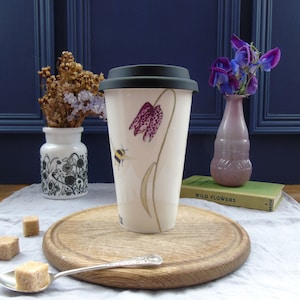 May include: A white ceramic travel mug with a black lid features a hand-painted illustration of a bee and a pink and white flower. The mug is sitting on a wooden chopping board.