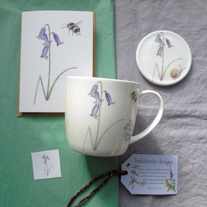 May include: A white mug with a bluebell and bee design. The mug is part of a set of bluebell themed gifts, including a card, coaster, and a small print.