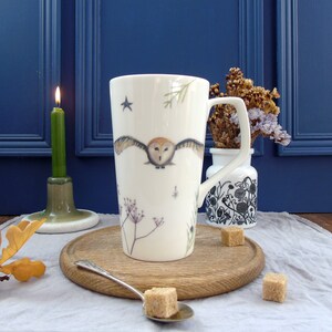 May include: A white ceramic mug with a brown owl design and a star. The mug is on a wooden coaster with a spoon and sugar cubes.