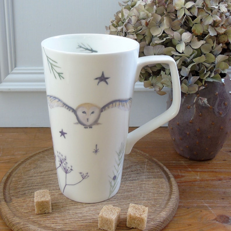 May include: A white ceramic mug with a tall, slender design features a watercolor illustration of a barn owl in flight with stars and sprigs of greenery.