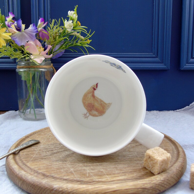 May include: A white ceramic mug with a handle, featuring a hand-painted illustration of a yellow chicken inside. The mug is sitting on a wooden surface.