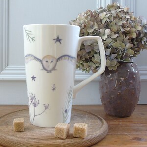 May include: White ceramic mug with a tall, slender shape and a large handle. The mug features a watercolor illustration of an owl in flight with stars and botanical elements.
