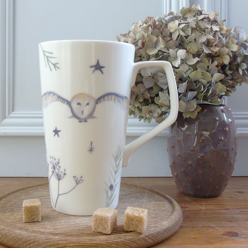 May include: White ceramic mug with a tall, slender shape and a large handle. The mug features a watercolor illustration of an owl in flight with stars and botanical elements.