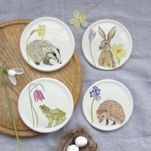 May include: Set of four white ceramic coasters with watercolour illustrations of a badger, a rabbit, a frog, and a hedgehog. Each coaster features a different wild animal and a flower.