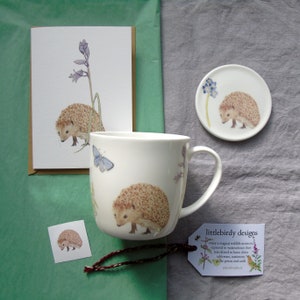 May include: A set of hedgehog themed gifts including a mug, a coaster, a card, and a sticker. The mug features a watercolor illustration of a hedgehog with butterflies and flowers. The coaster is a round ceramic piece with a watercolor illustration of a hedgehog and blue flowers. The card is a white card with a watercolor illustration of a hedgehog and blue flowers. The sticker is a small square sticker with a watercolor illustration of a hedgehog.