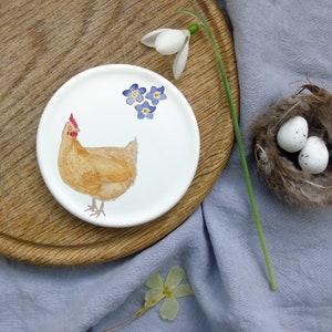May include: A white ceramic dish with a hand-painted illustration of a brown chicken and blue forget-me-not flowers. The dish is on a wooden surface.